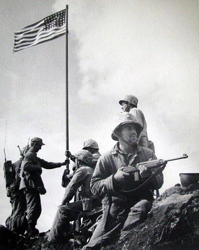 This is Marine photographer Lou Lowery's picture of the first flag-raising. In the foreground holding a carbine is Pfc. James Michels and behind him is Cpl. Charles Lindberg. Lt. Harold Schrier, holds the flag pole on the left, and behind him is Pfc. Louis Chario, hardly visible with a radio on his back. Sgt. Harry Hansen holds the flag pole from the right while Sgt. Ernest Thomas looks on. Photo provided by Dick Honyak