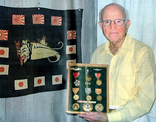 Capt. Ralph Styles, former skipper of the USS Sea Devil, holds a shadow box full of medals including two Navy Crosses, two Legions of Merit, Navy Unit Commendation, a solid gold submarine pin for being awarded two Navy Crosses. In the background is a banner with the USS Sea Devil's emblem surrounded by five Japanese rising sun flags indicating five enemy war ships sunk or damaged and seven white flags with red centers denoting seven enemy transports sunk. Sun photo by Don Moore