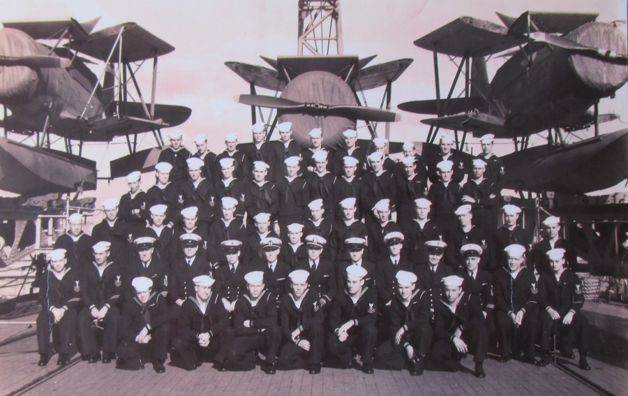 The engine-room crew of the USS Philadelphia on its fantail during the Second World War. Note the three covered patrol planes with wings folded flanking the group. Photo provided 